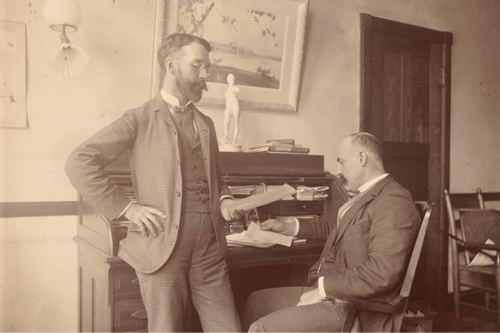 Two men are around a wooden desk. One (Charles M. Kurtz) is standing. He is bearded and wearing a checkered suit. The other (Halsey C. Ives) is seated. He has a mustache and is wearing a dark suit. 